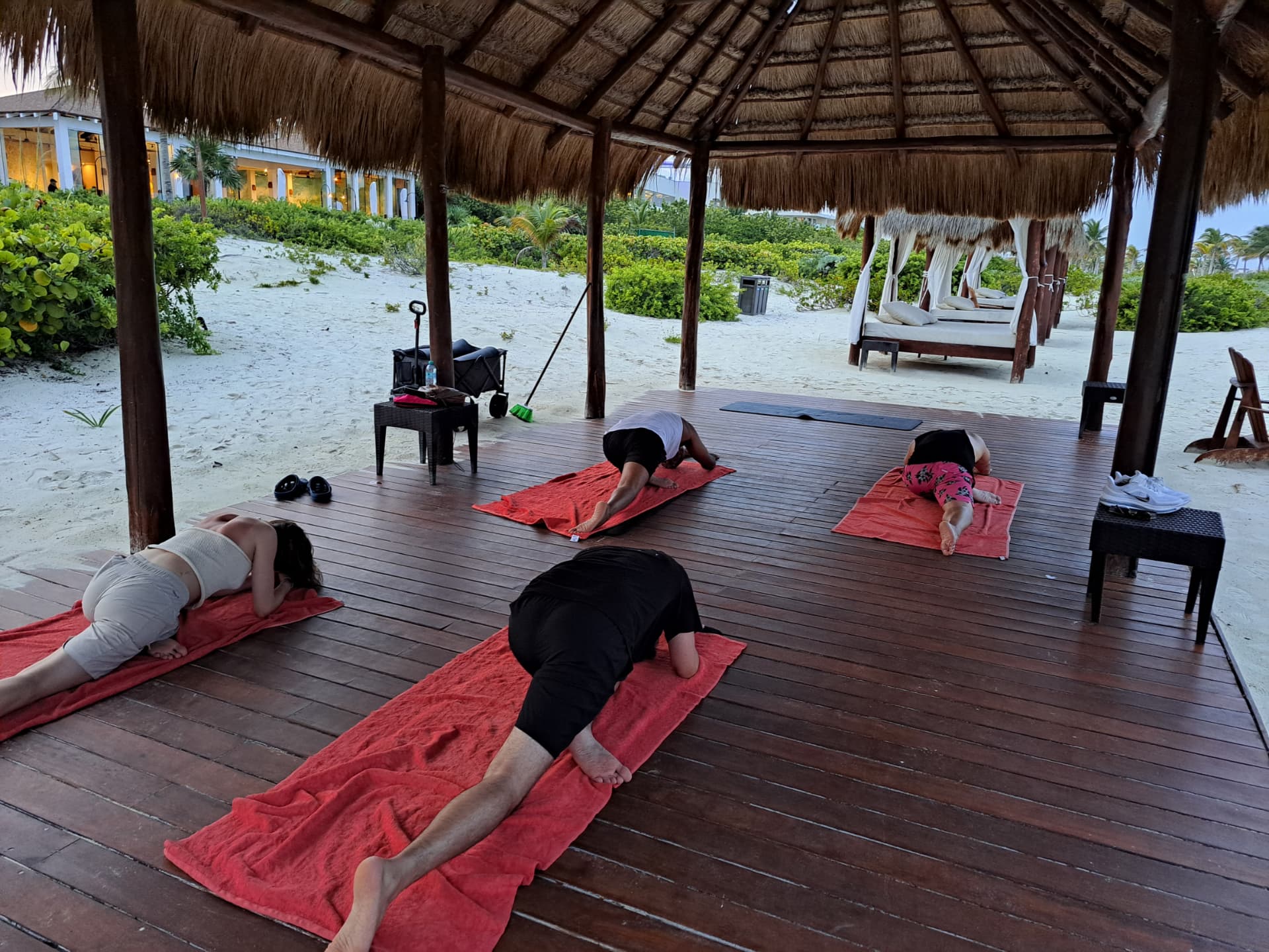 Group yoga class holding a hip-opening stretch on a wooden deck under a thatched-roof palapa at a beach resort