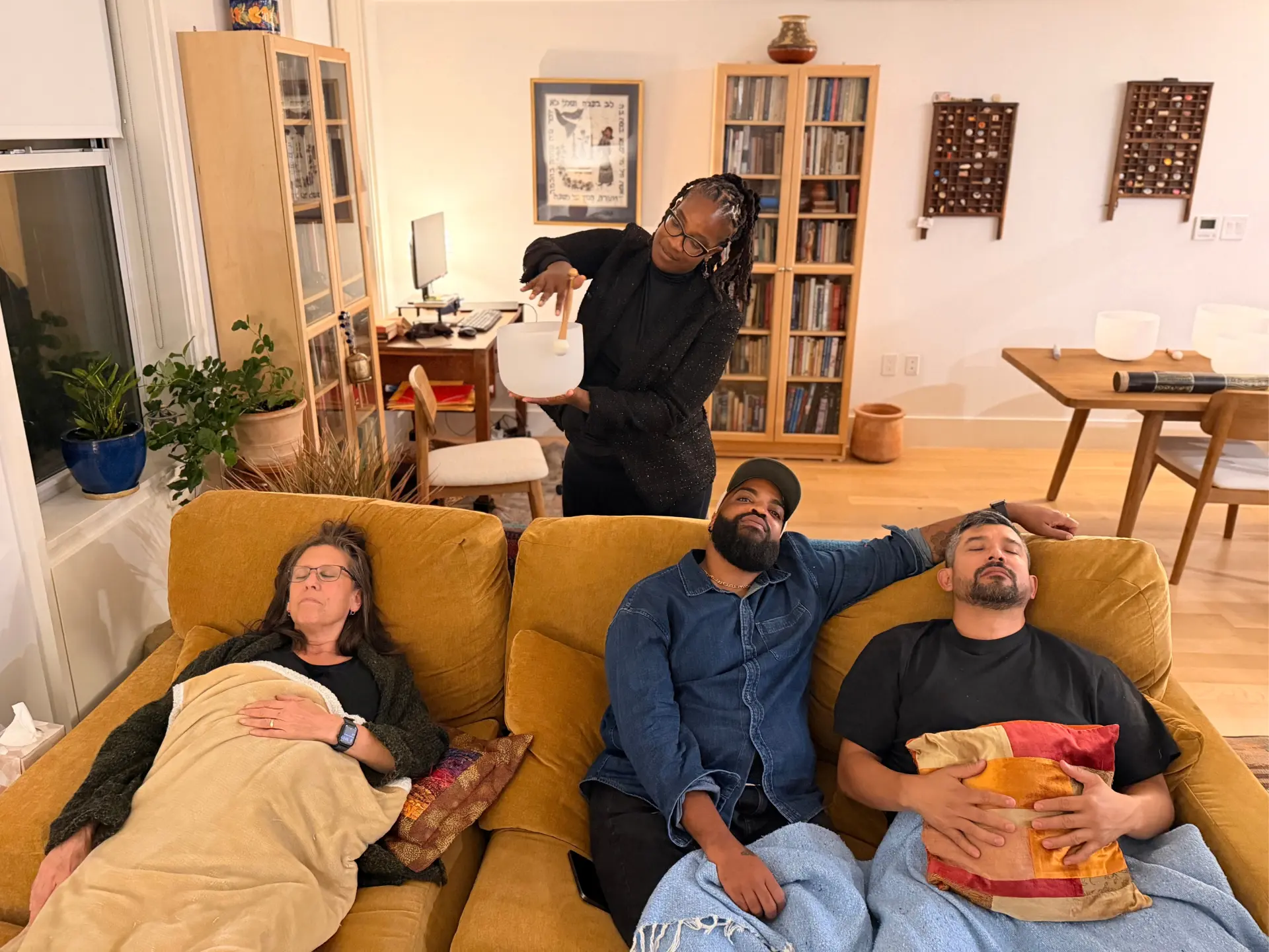 Keita playing a crystal singing bowl behind a mustard-yellow couch while three guests rest with eyes closed in a sunlit living room
